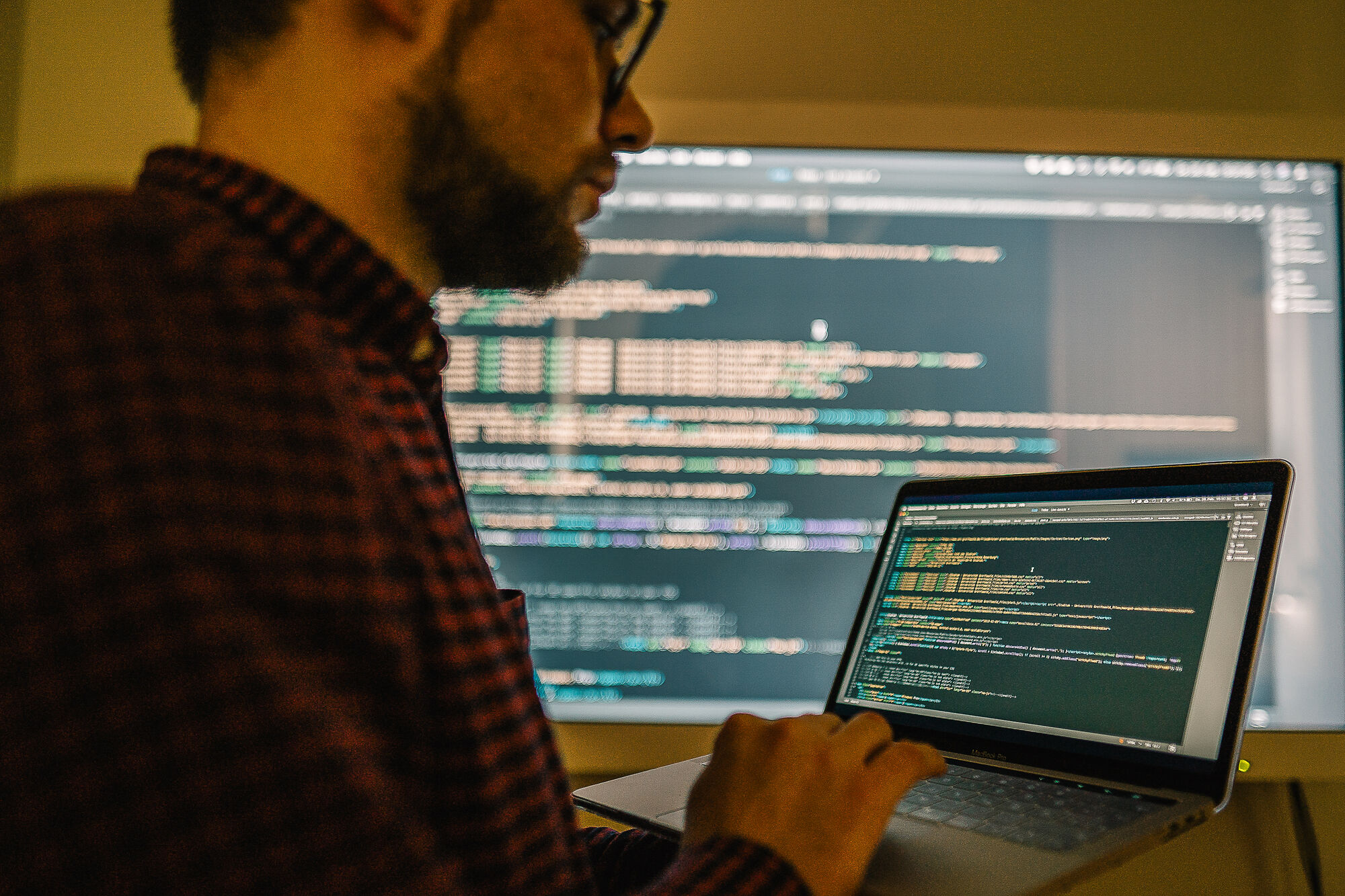 Ein Mann bearbeitet auf einem Notebook einen Programmiercode, der auf einem beleuchteten Bildschirm ausgespielt wird.