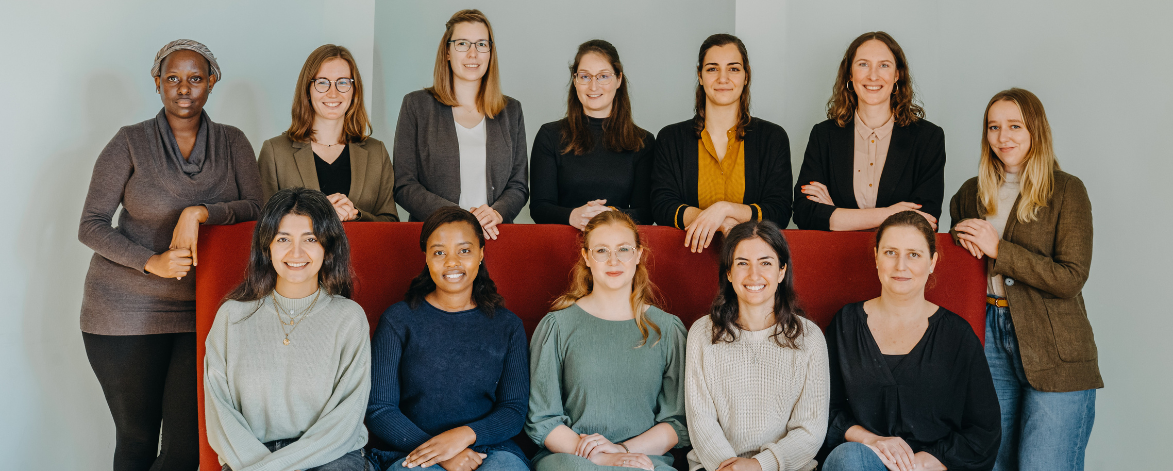 12 Frauen stehen vor einer hellen Wand. 5 davon sitzen auf einem roten Sofa, die anderen 7 stehen dahinter. Alle schauen in die Kamera.