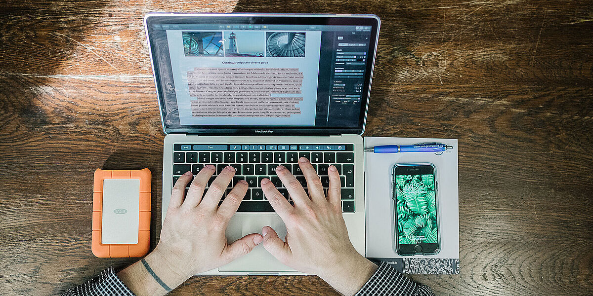 Jemand arbeitet am Laptop. Daneben liegt ein Schreibblock, ein Stift sowie ein Smartphone.