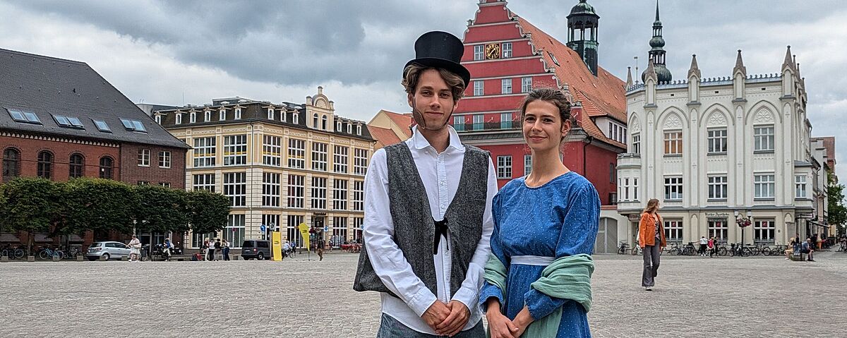 Anlässlich des Caspar-David-Friedrich-Jahres 2024 stehen ein Mann und eine Frau in historischer Tracht auf dem Marktplatz in Greifswald.