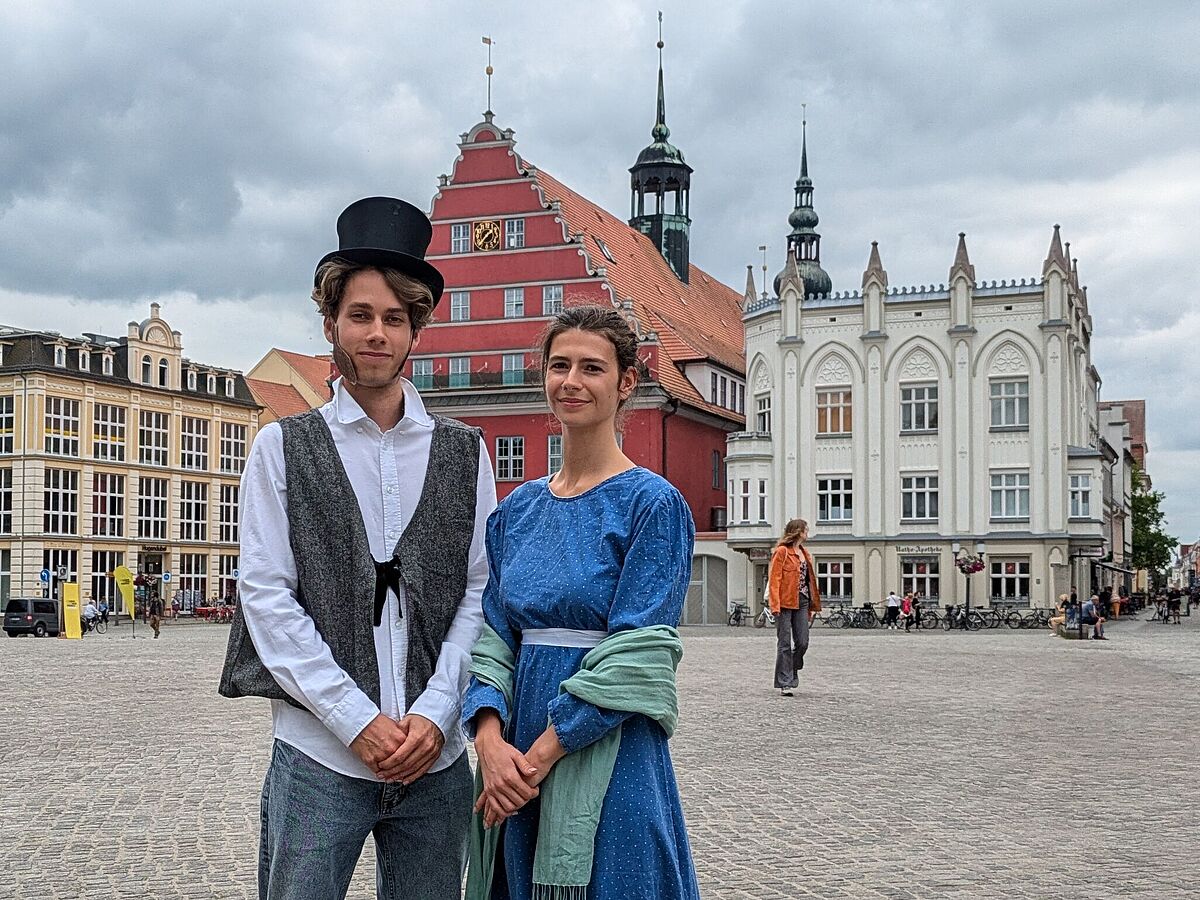 Anlässlich des Caspar-David-Friedrich-Jahres 2024 stehen ein Mann und eine Frau in historischer Tracht auf dem Marktplatz in Greifswald.