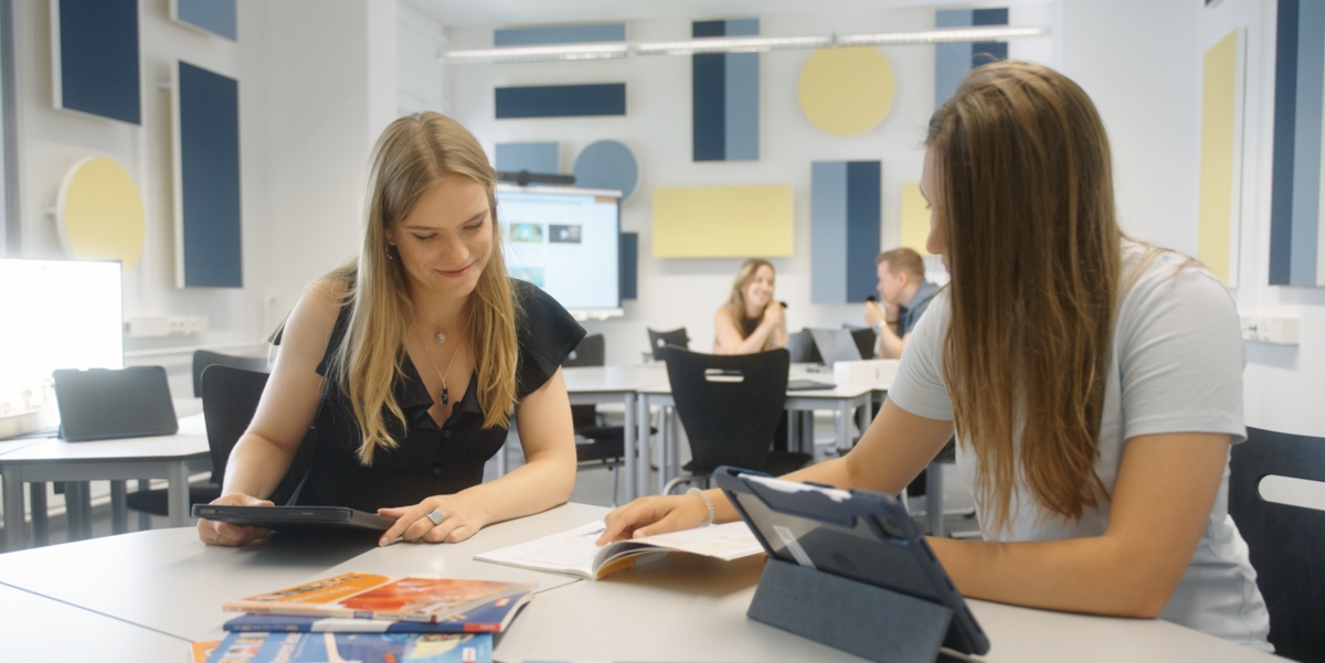 Zwei Studierende sitzen an einem Tisch, vor ihnen ist ein Tablet und verschiedene Arbeitshefte. 