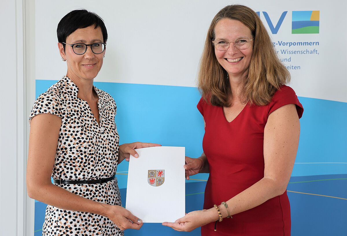 Auf dem Bild sehen wir, wie Wissenschaftsministerin Bettina Martin die Ernennungsurkunde an Dr. Juliane Huwe übergibt. Beide halten die Urkunde in die Kamera. 