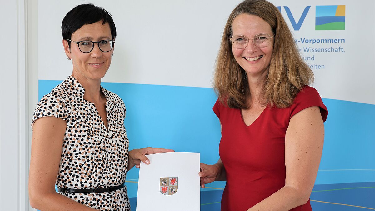 The image depicts Minister of Science Bettina Martin handing over the certificate of appointment to Dr. Juliane Huwe. Both hold the certificate to the camera.