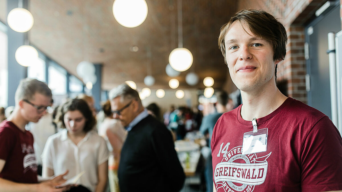 Viele Personen informieren sich bei den Hochschulinfotagen über Studienmöglichkeiten in Greifswald. Ein Campusspezialist schaut freundlich in die Kamera.