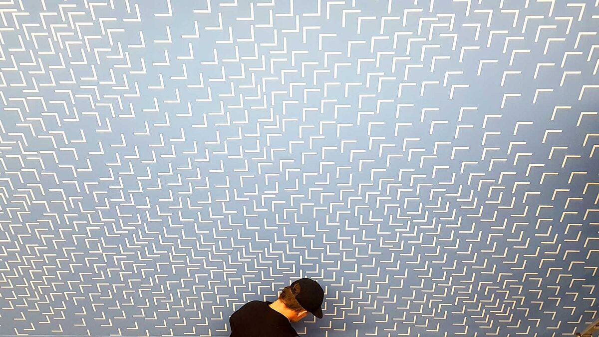 Man sieht ein Kunstwerk von Susanne Gabler, ein großes Bild mit vielen weißen rechten Winkeln auf hellblauem Hintergrund. Sie sind nicht ganz regelmäßig verteilt und in unterschiedliche Richtungen gedreht, sodass ein 3D-Täuschung entsteht.Davor der Kopf einer besuchenden Person, die das Werk betrachtet.