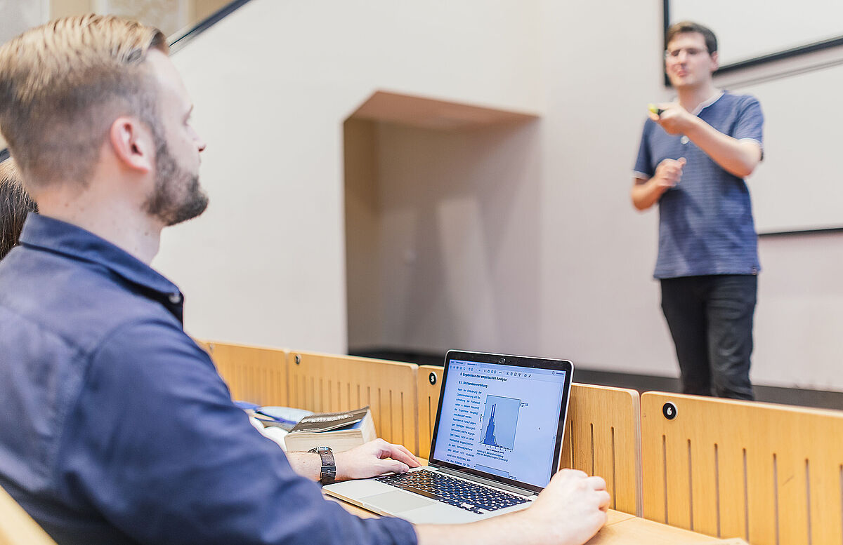 Ein Mann sitzt in einem Hörsaal vor einem Laptop mit geöffnetem Diagramm, während ein Dozent vorne spricht und gestikuliert.