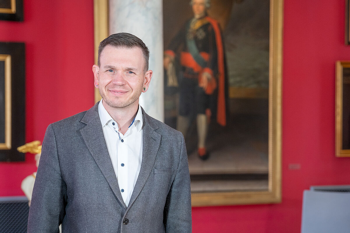 Das Bild zeigt Dr. Michael Schöner, den Leiter der Graduiertenakademie, vor dem Hintergrund eines Gemäldes in der Aula der Universität. Er trägt ein weißes Hemd und graues Sakko.