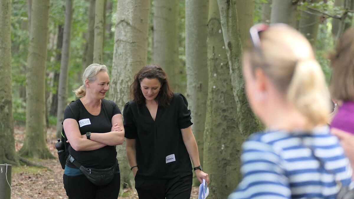 Zwei Frauen laufen nebeneinander durch den Wald und unterhalten sich. Im Vordergrund sind weitere Frauen unscharf zu sehen.
