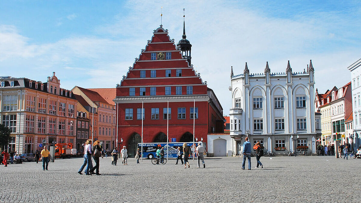 Blick über den Greifswalder Markt auf das Rathaus der Stadt. Es ist schönes Wetter und Leute gehen über den Markt.