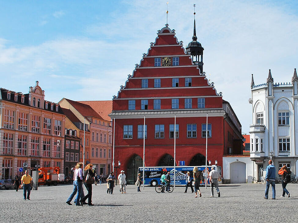 Blick über den Greifswalder Markt auf das Rathaus der Stadt. Es ist schönes Wetter und Leute gehen über den Markt.