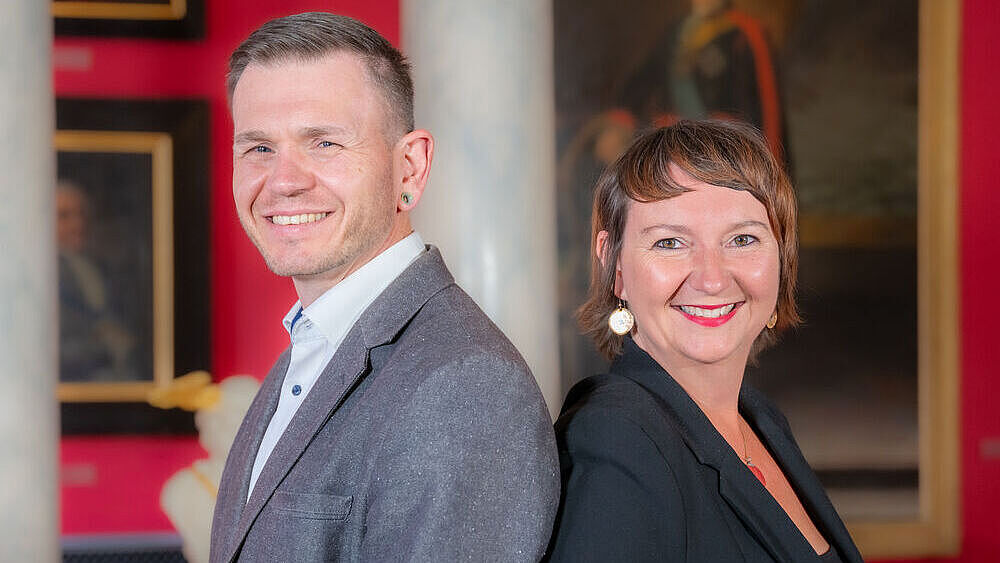 Auf dem Bild sind Dr. Jana Kiesendahl und Dr. Michael Schöner zusammen zu sehen. Sie stehen in der Aula der Universität. 