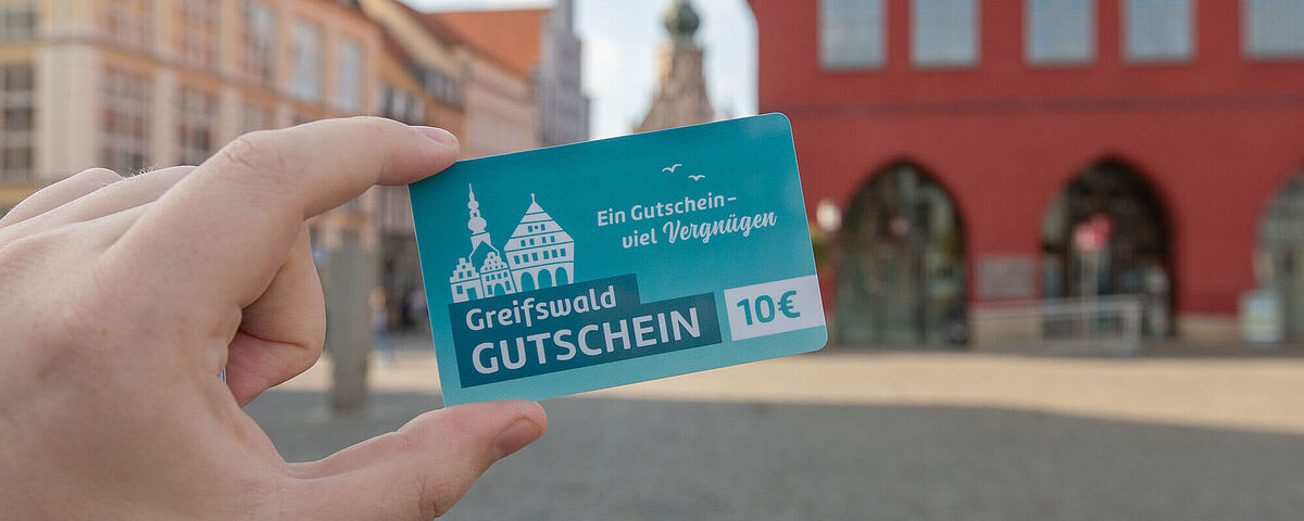 Eine Person auf dem Marktplatz hält einen hellblauen Greifswald-Gutschein in Richtung Rathaus.-