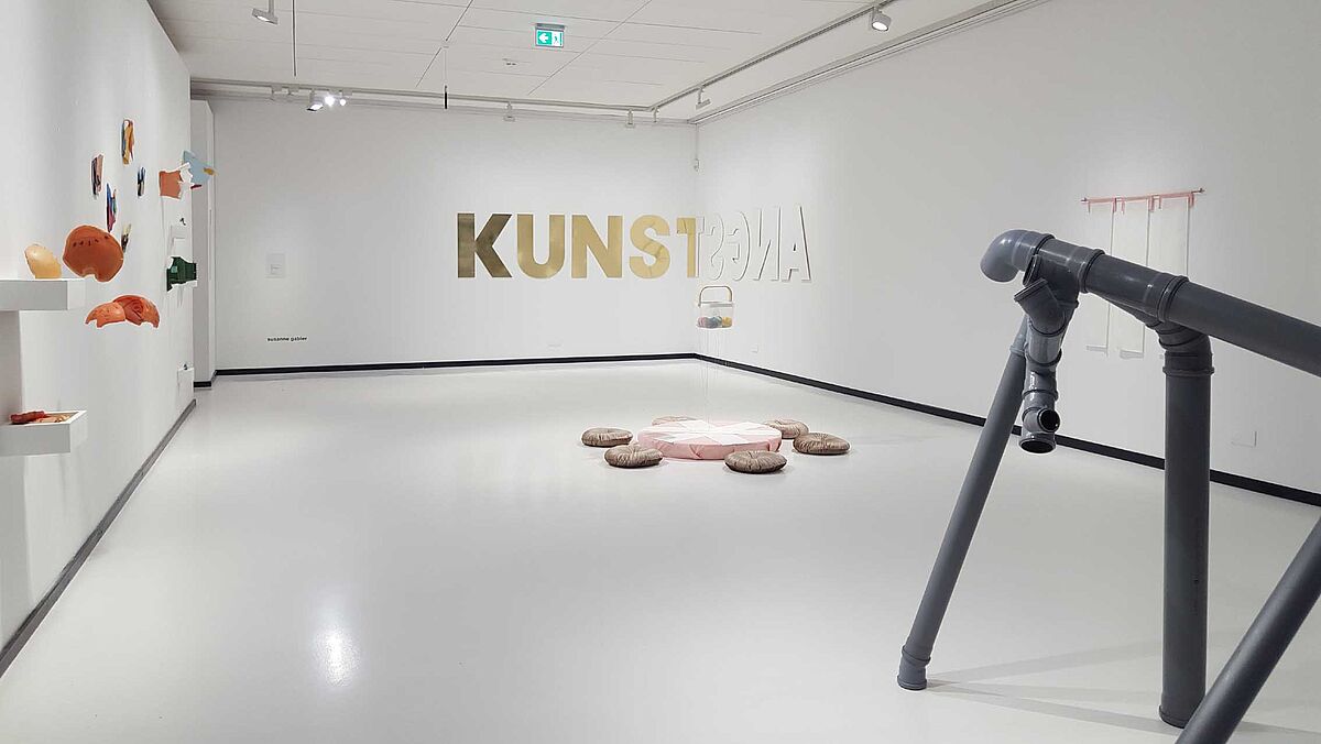 Man sieht einen Ausstellungsraum mit verschiedenen Kunstwerken von Susanne Gabler, unter anderem HT-Röhren, die zusammengesetckt wurden, sodass sie auf dem Boden stehen wie ein Gerüst. Auch das Kunstwerk "KUNSTSGNA" ist in der Raumecke zu sehen. Dieses wird im nächsten Bild beschrieben. 