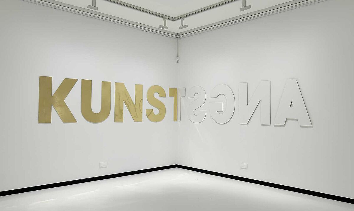Man sieht ein Kunstwerk von Susanne Gabler. An einer Wand sind große Buchstaben zu sehen.zur Raumecke hin steht "KUNST", an der andere Wand, zur selben Raumecke verlaufend steht von rechts nach links "ANGST". Jeweils das "T" am Ende des Wortes ist der selbe Buchstabe genau in der Raumecke. "KUNST" ist gold, "Angst" weiß geschrieben.